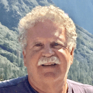 Smiling older man with curly gray hair and mustache, outdoors with mountains and trees in the background.