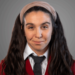 Woman with long dark hair in pigtails, wearing a red blazer, white shirt, black tie, and beige headband, smiling.