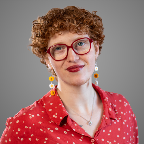 Person with short curly red hair, red glasses, floral earrings, and a red patterned shirt, smiling slightly.
