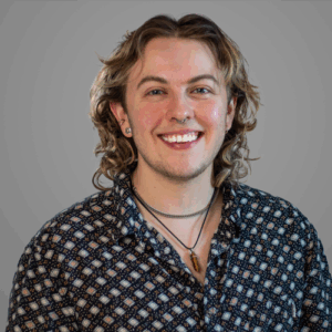 Smiling person with wavy hair, wearing patterned shirt and necklaces, in front of a plain gray background.
