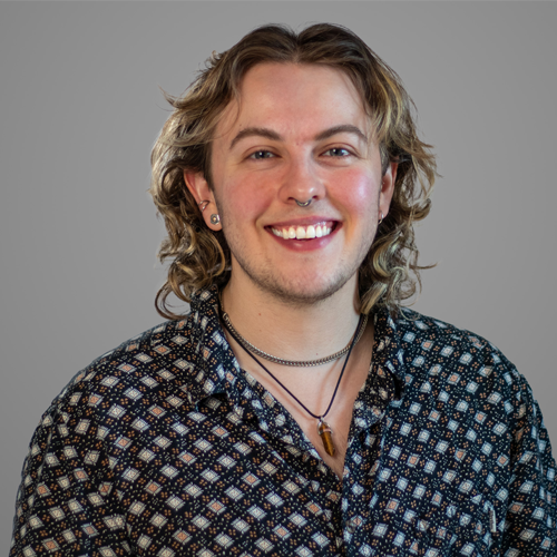 Smiling person with wavy hair, wearing patterned shirt and necklaces, in front of a plain gray background.