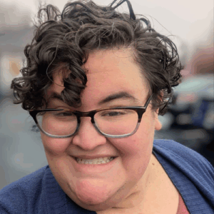 Person with short curly hair, glasses, and a blue sweater smiling outdoors on a cloudy day.
