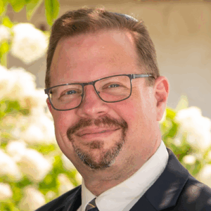 Smiling man with glasses and a goatee, wearing a suit and tie, outdoors with greenery in the background.