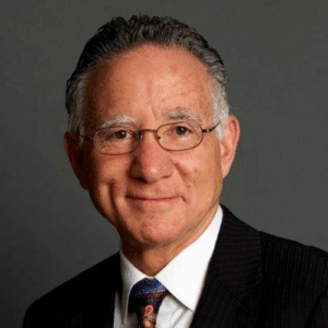 Smiling older man with glasses, gray hair, and a suit against a plain gray background.