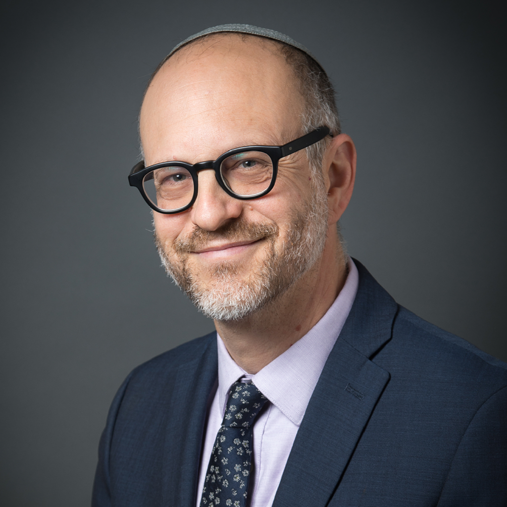 Smiling man in glasses, suit, and tie, wearing a kippah, posed against a plain dark background.