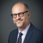 Smiling man in glasses, suit, and tie, wearing a kippah, posed against a plain dark background.