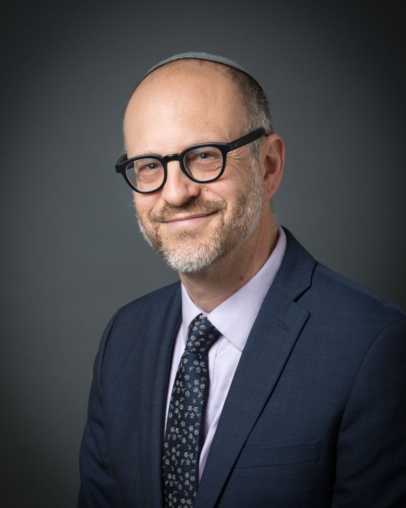 Smiling man in glasses and a suit jacket with a patterned tie, posing against a dark gray background.