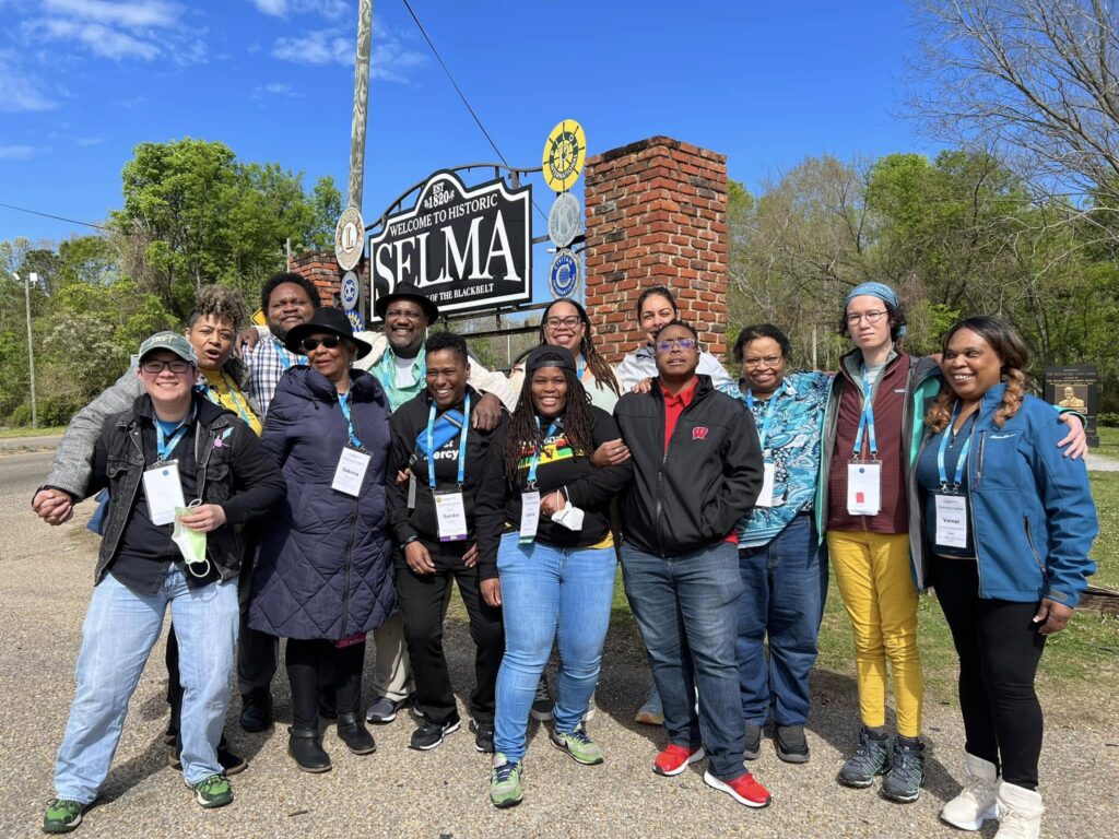 A diverse group poses and smiles in front of the Welcome to Historic Selma sign on a sunny day.