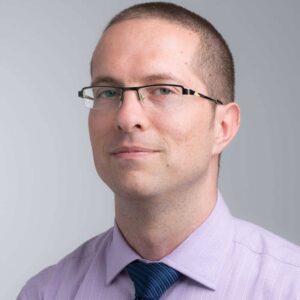 Man with short hair, glasses, and a light purple shirt with a blue tie, looking at the camera.