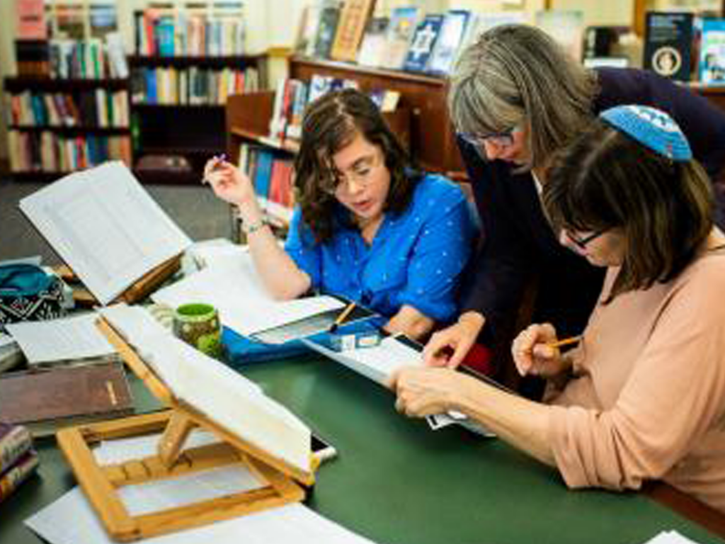 Three women study together at a table with books and papers in a library or study room.