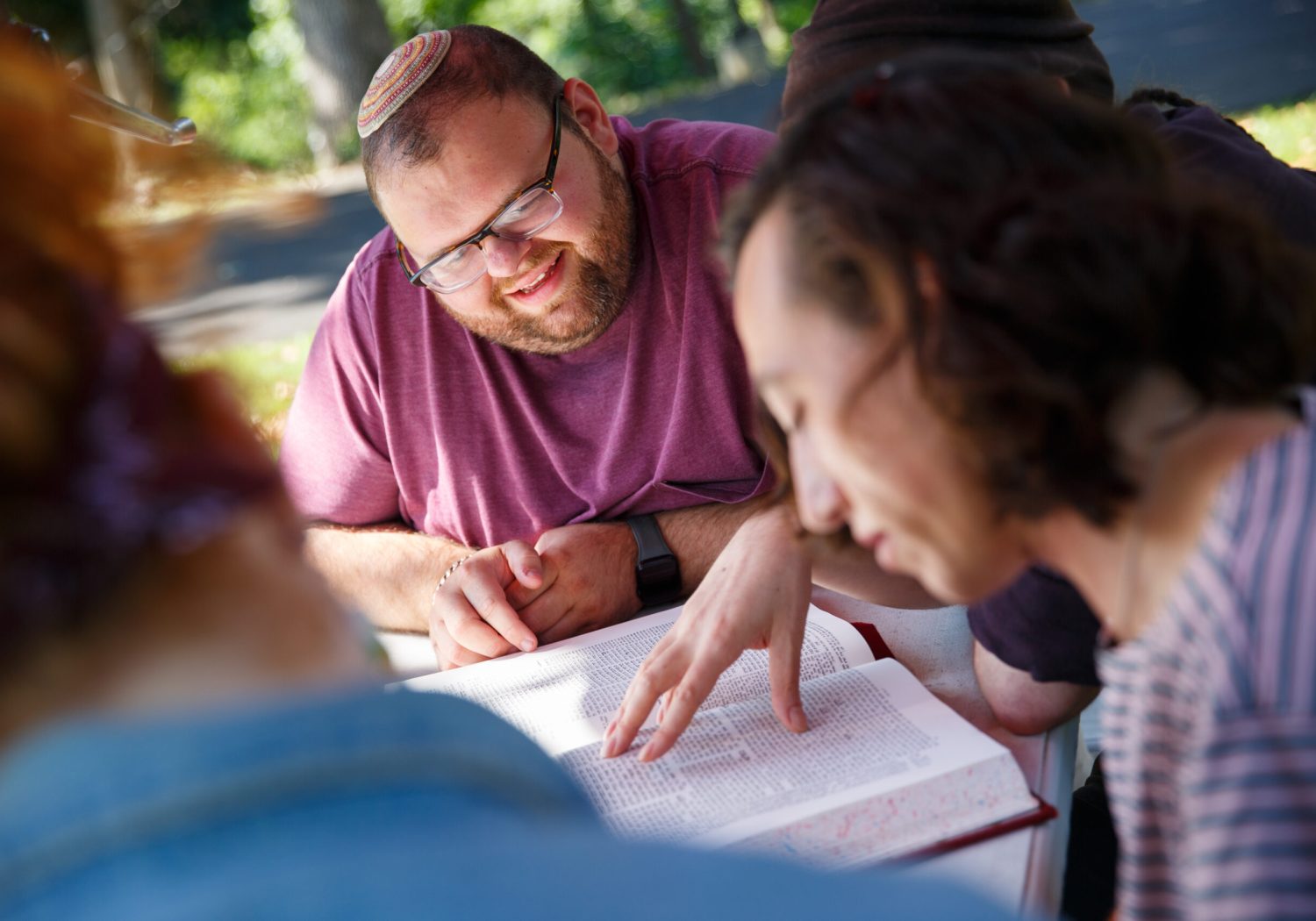A group of people studies a book outdoors, one person points at the text while others look on.