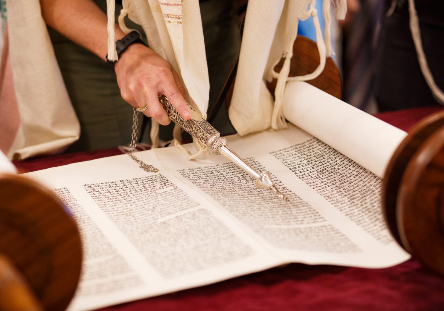 A person uses a silver yad pointer to read from an open Torah scroll on a table.